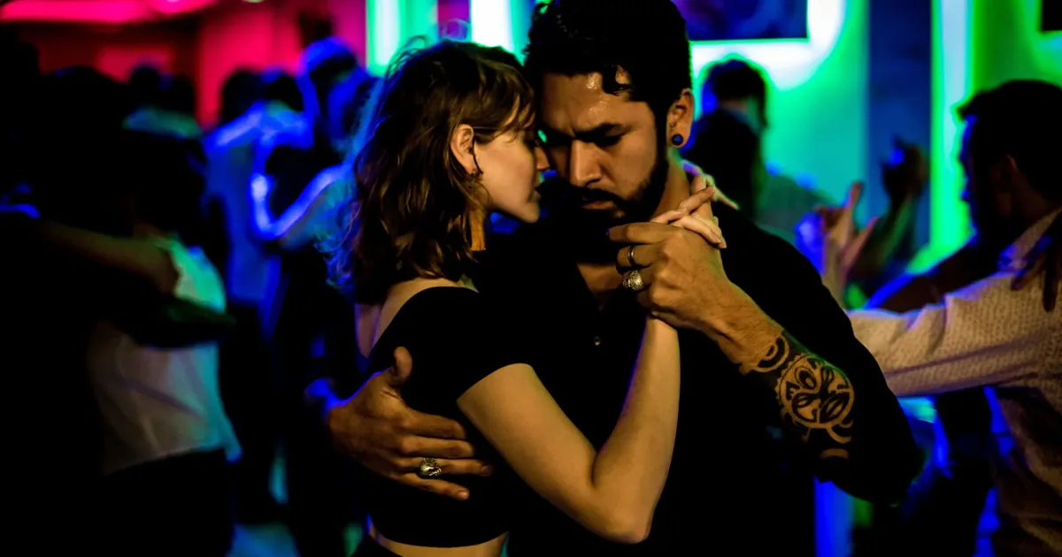 A couple in close embrace on a crowded Buenos Aires milonga floor, the kind of social dancing a beginner class prepares you for