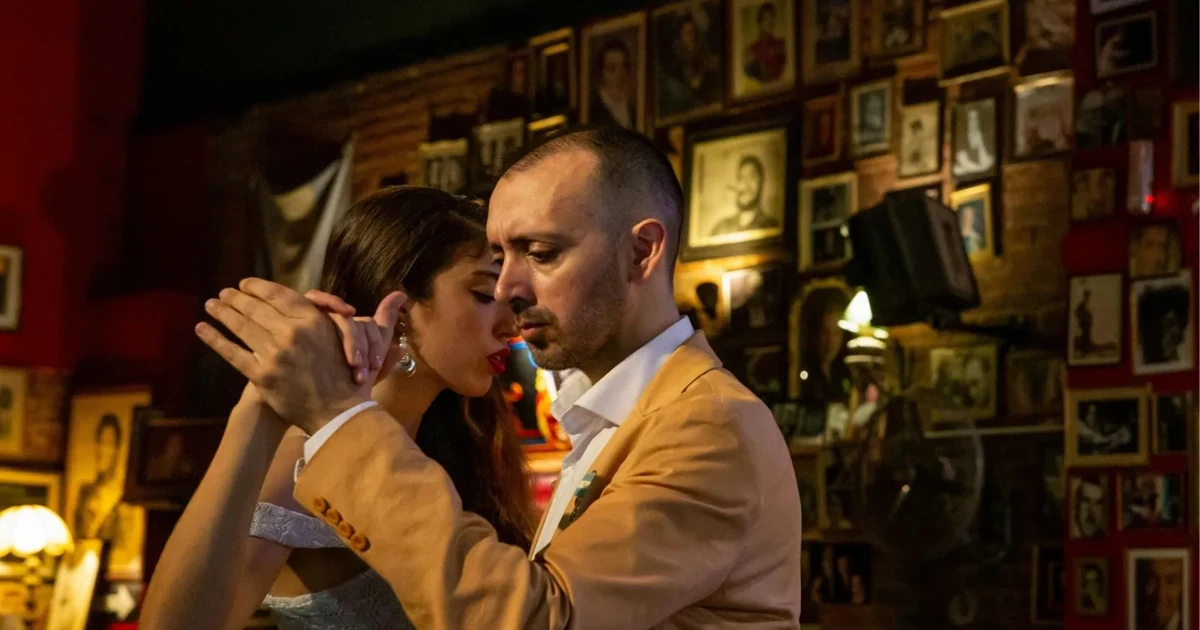 A couple dancing tango in a historic Buenos Aires café whose walls are covered floor to ceiling with framed photographs, set for dinner