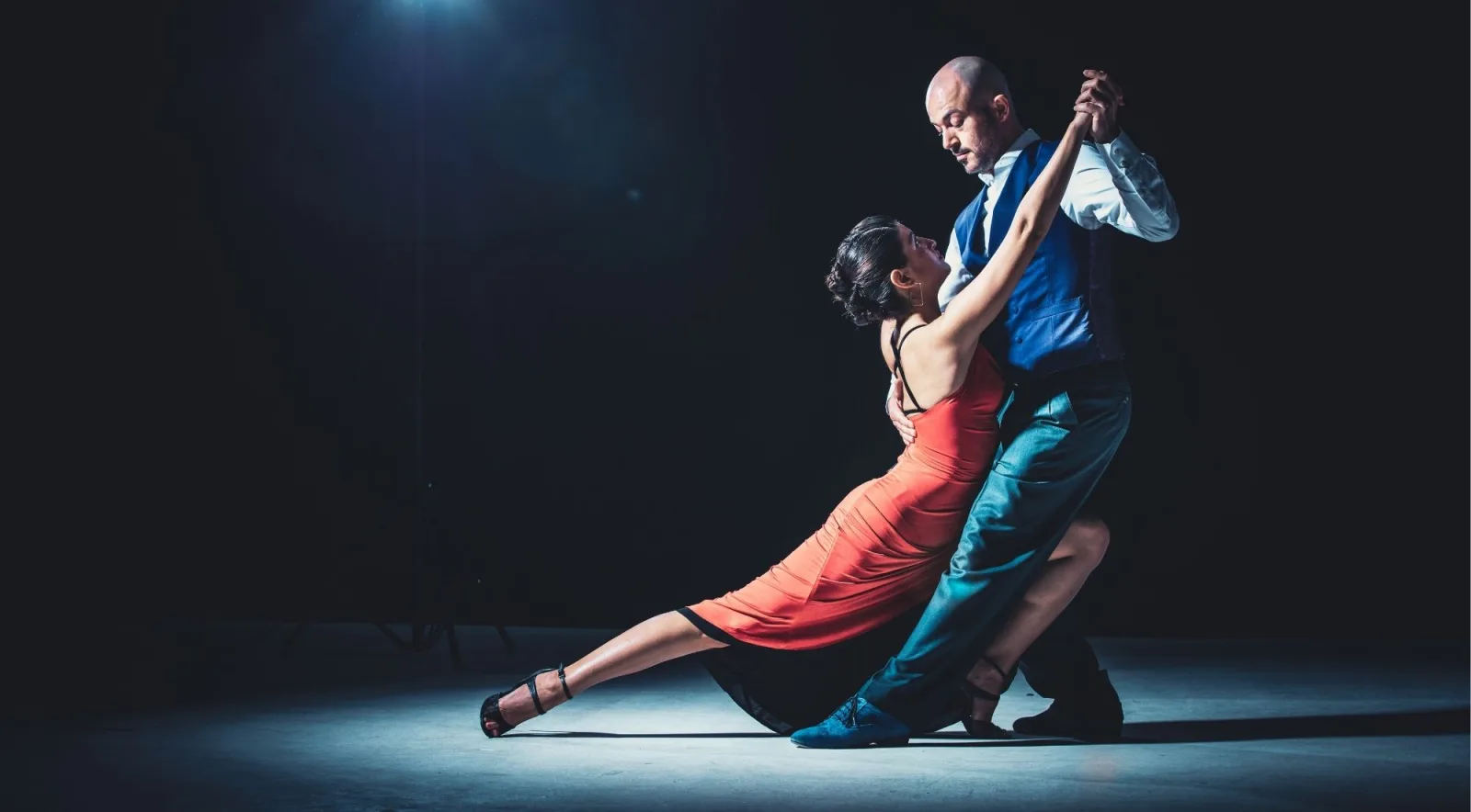 Tango couple in a deep lunge under a single spotlight, the woman in a red dress, the man in a blue waistcoat
