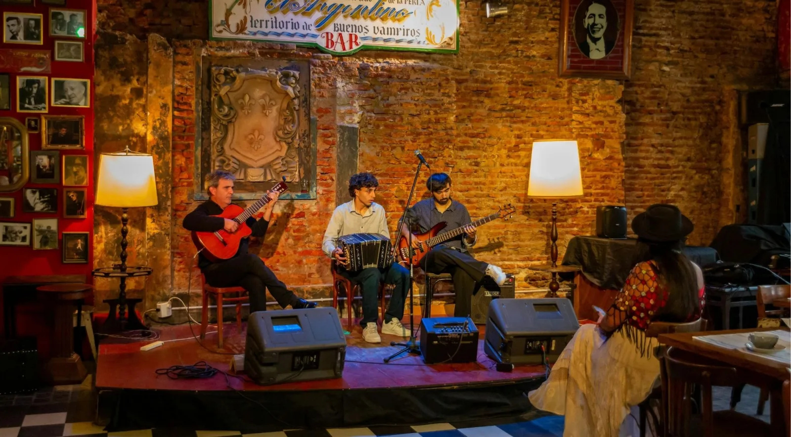 A three-piece tango band playing guitar and bandoneón on a small stage in a brick-walled bar hung with framed photographs