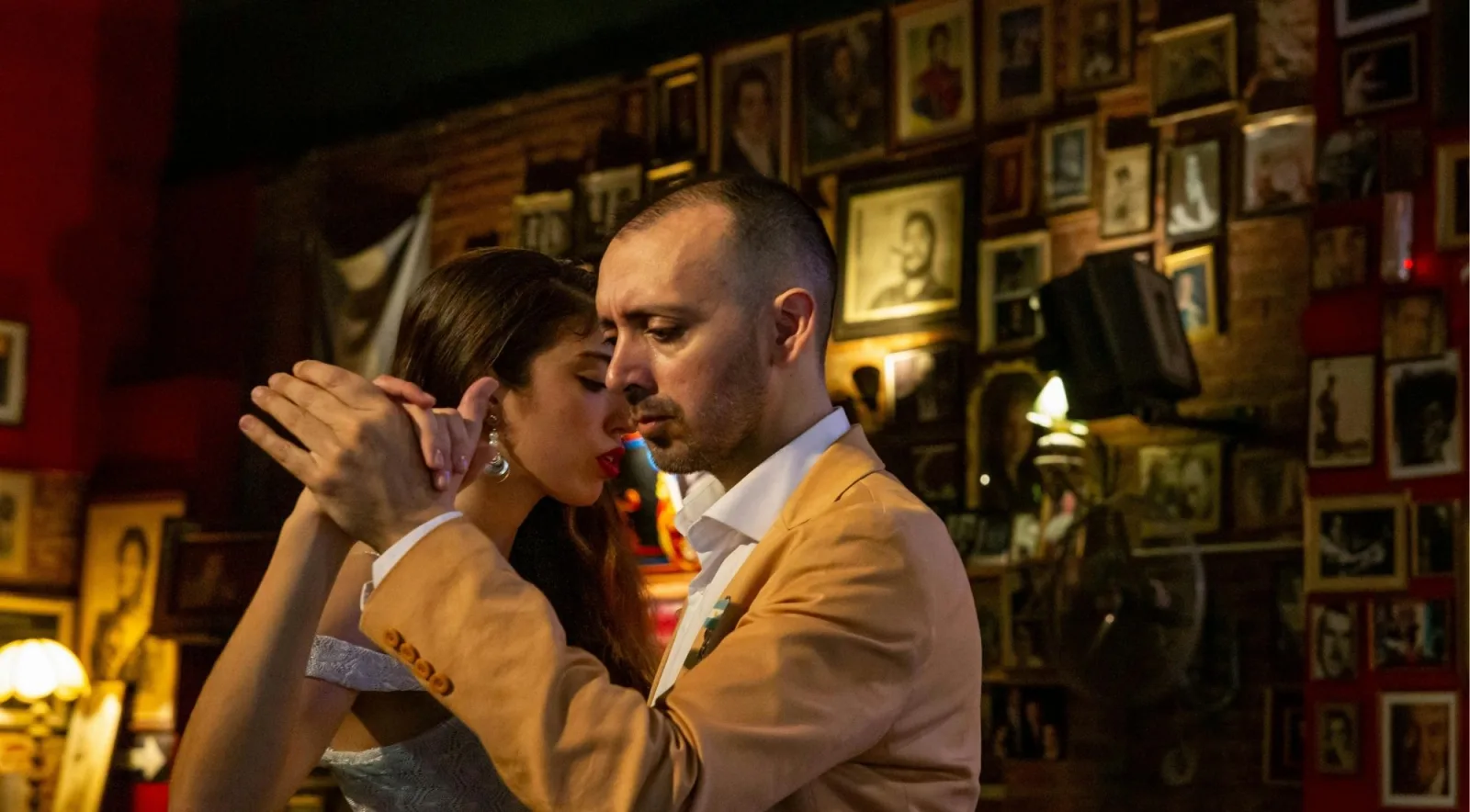 A couple dancing cheek to cheek in a tango bar whose walls are covered floor to ceiling with framed portraits