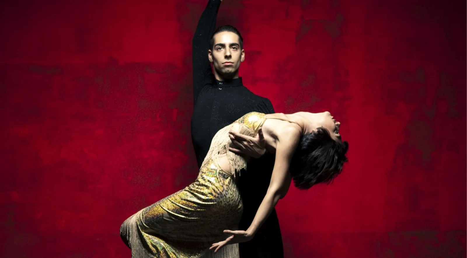 Staged tango performance against a deep red wall, the woman arched backwards in a gold dress