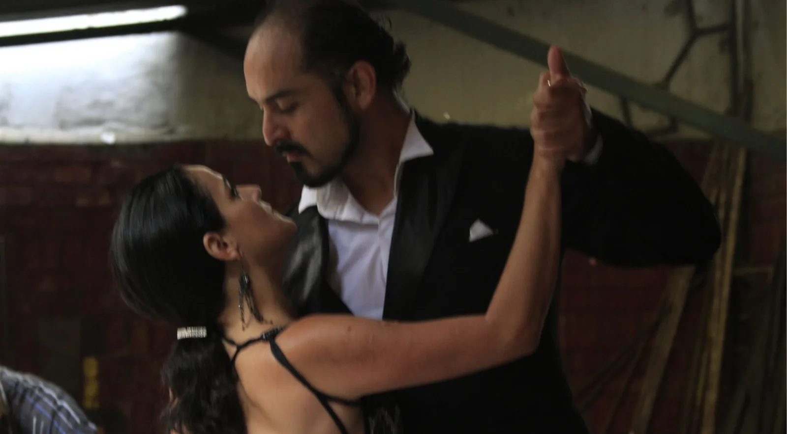A couple in close embrace, the man in a black tuxedo, lit from one side against a dark wall