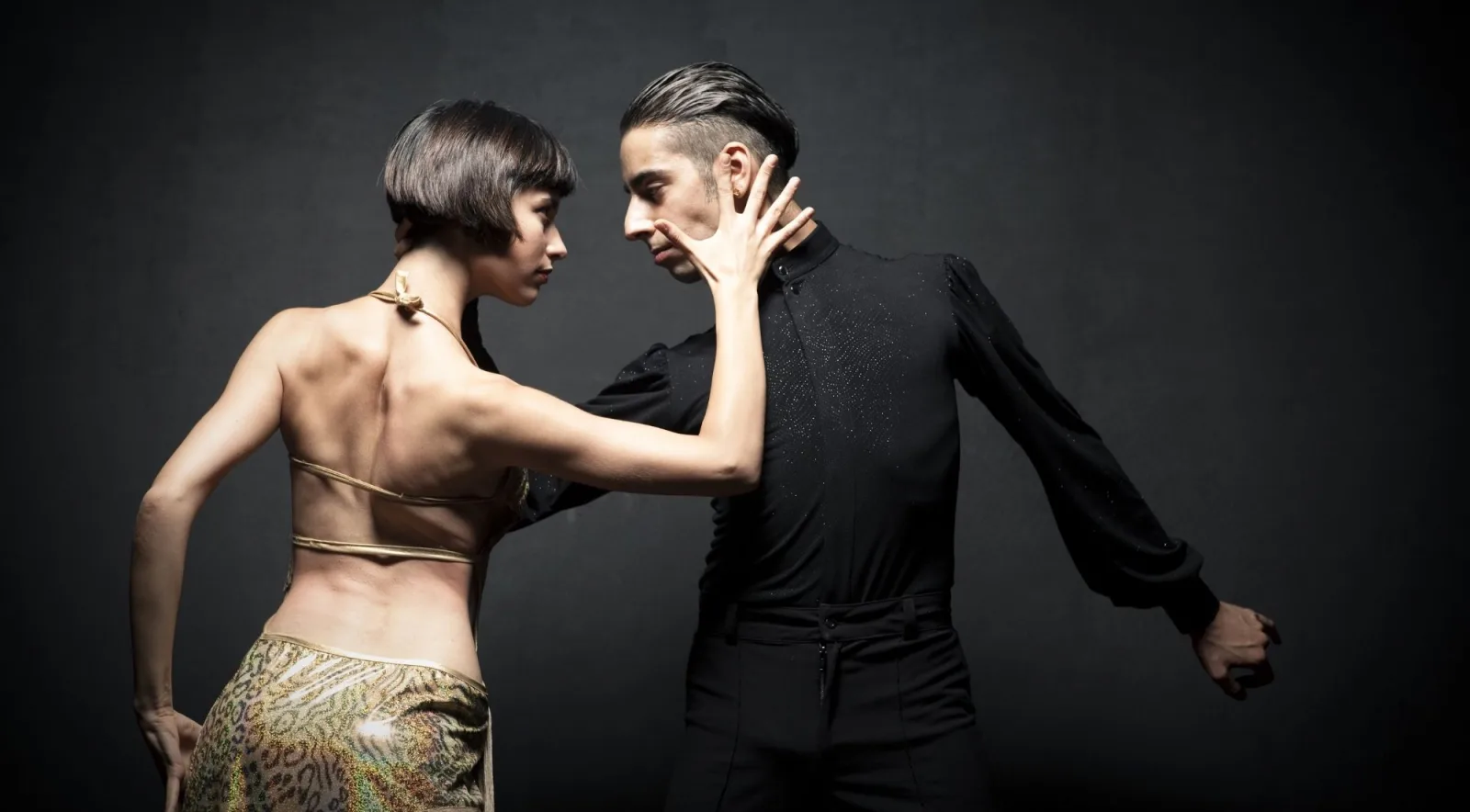 Studio portrait of a tango couple in profile, the woman in a gold patterned dress, against a grey backdrop