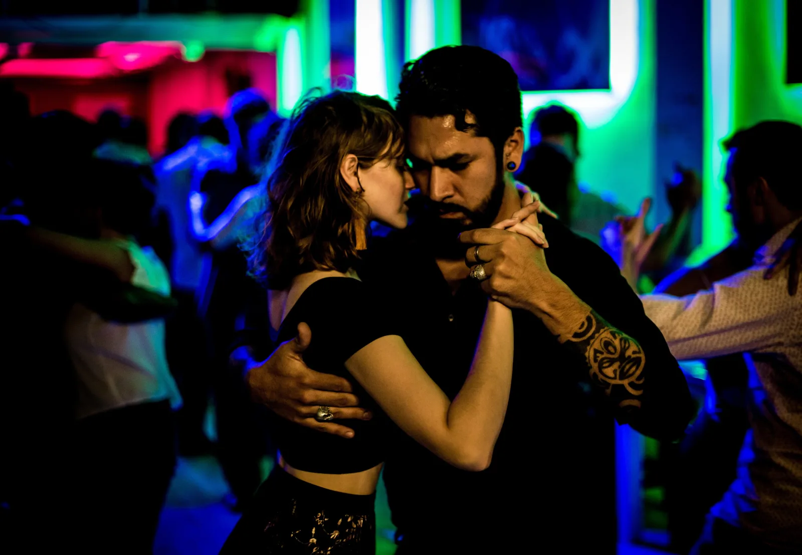 A couple in close tango embrace on a milonga floor lit in green and blue