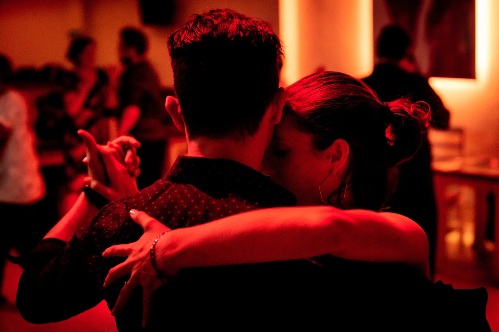 A tango couple dancing in warm red light among other dancers at a milonga