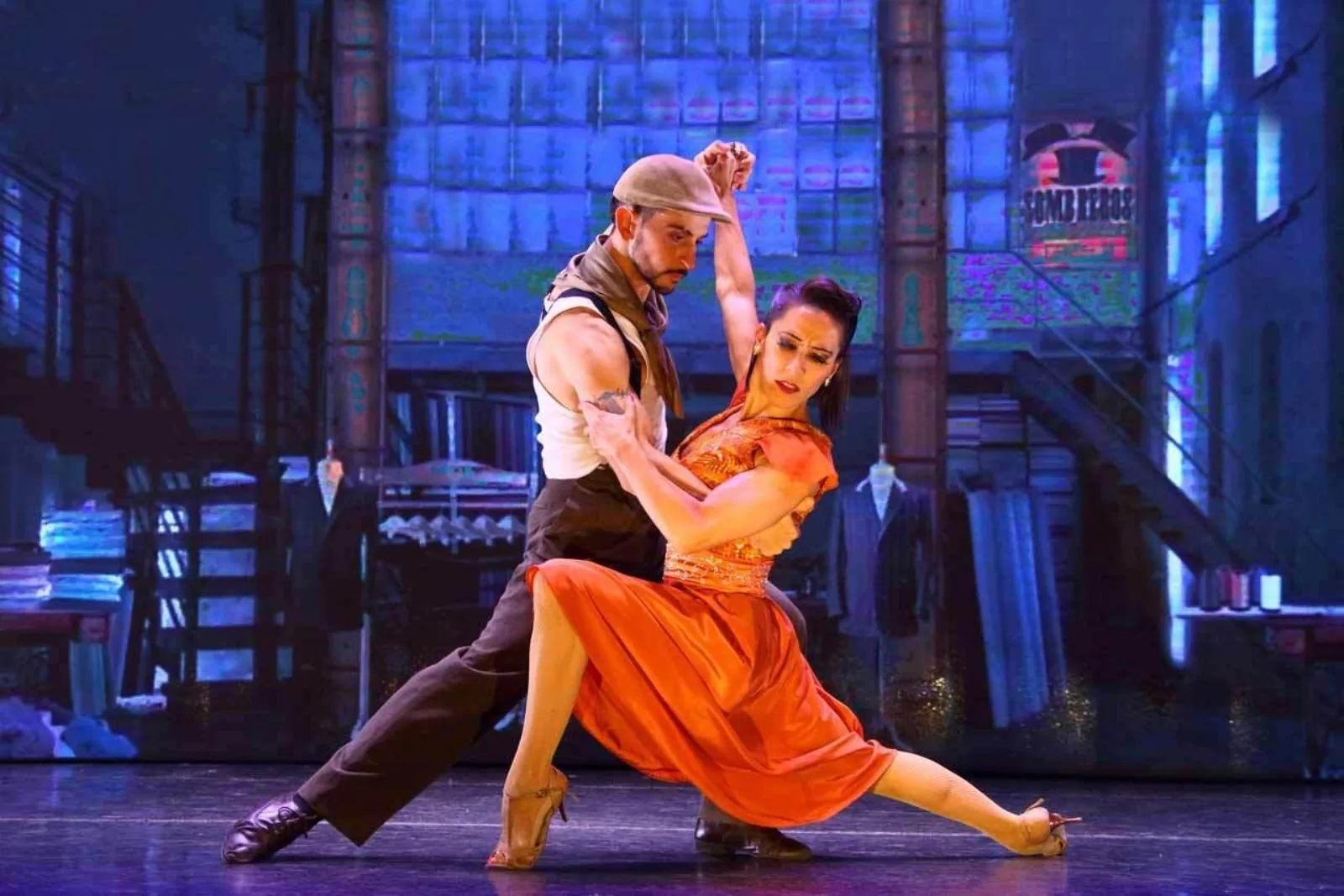 Two Madero Tango dancers mid-step in orange costume against a painted street backdrop